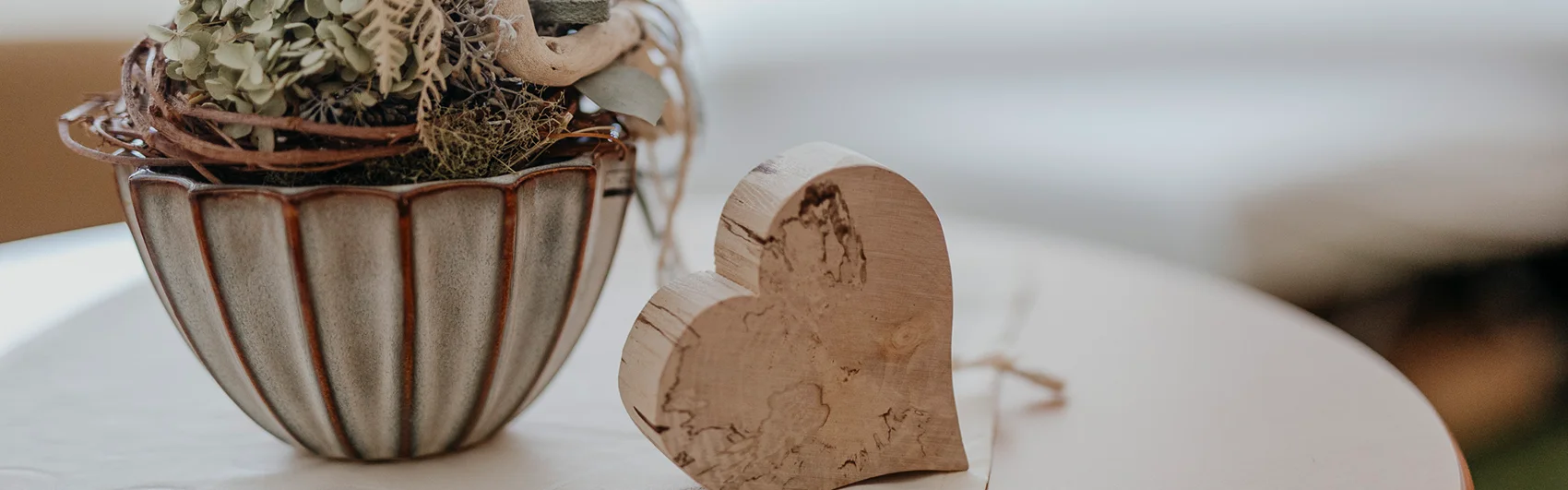 Im Vordergrund ein runder Tisch mit einem Holzherz und einem kleinen Blumenstock, im Hintergrund das frisch gemachte Bett.